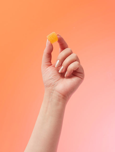 Lifestyle image of a hand holding a gummy, orange and pink background